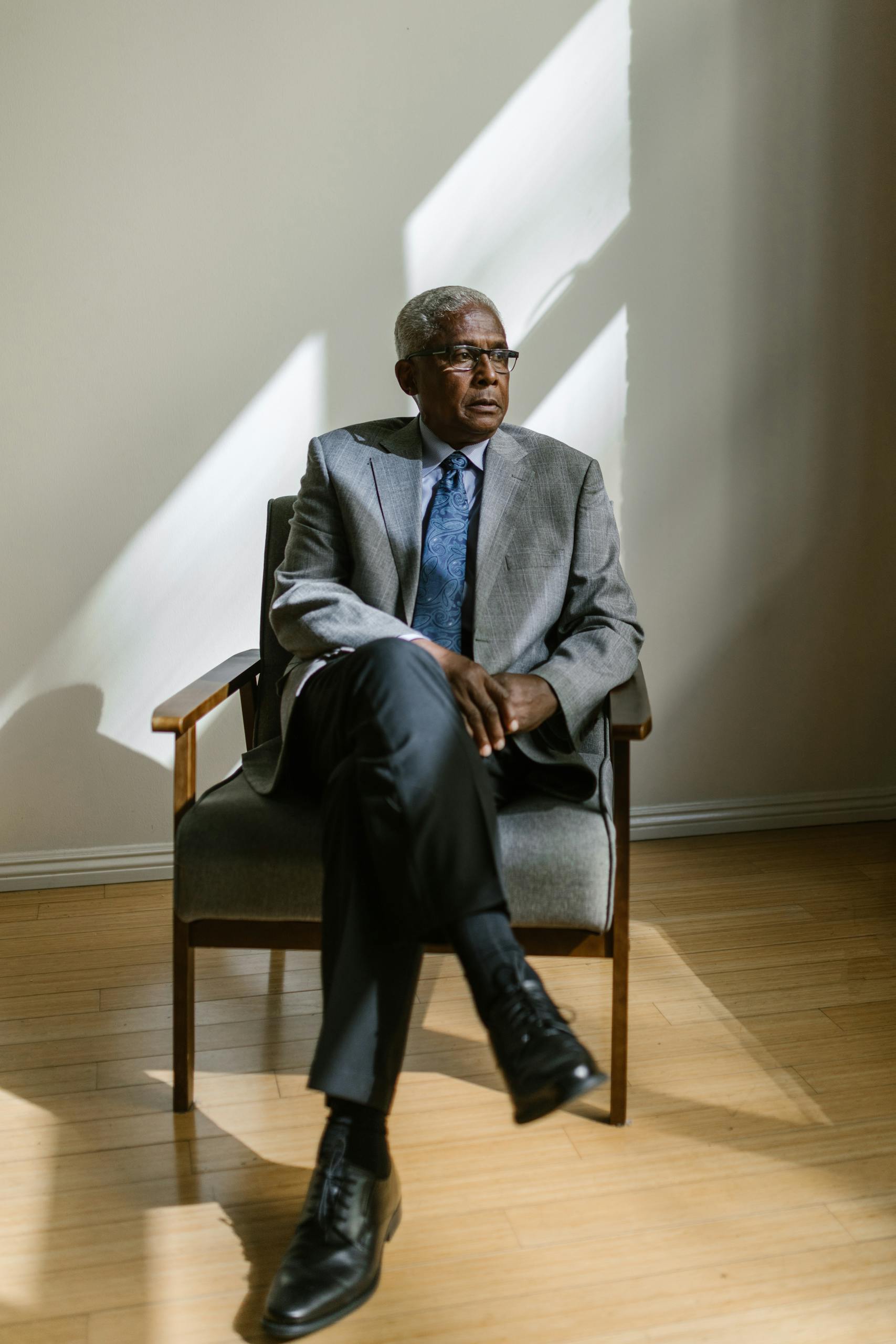 Senior man in a gray suit sitting in a wooden armchair in a well-lit room.